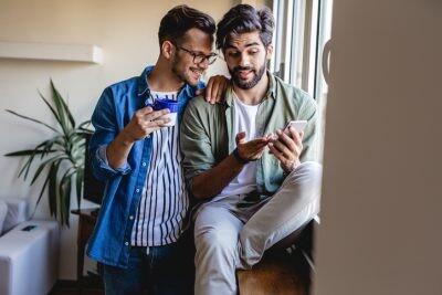 Gay men couple at home sit by the window and look at screen of the smartphone for a hook up