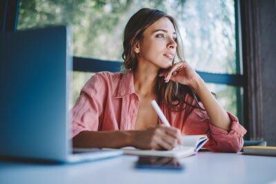 Pondering young woman looking away while making notes and thinking on creative ideas for developing own internet website.Thoughtful hipster student writing down information in notepad sitting at table looking at dealbreakers