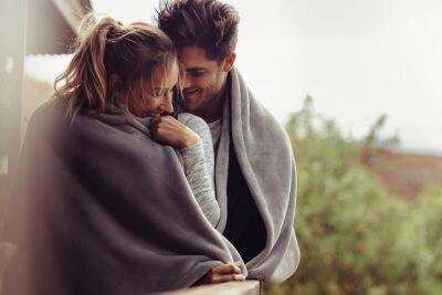 Romantic couple on a winter holiday. Man and woman standing together in a hotel room balcony wrapped in blanket. Couple embracing and smiling during the holiday season.